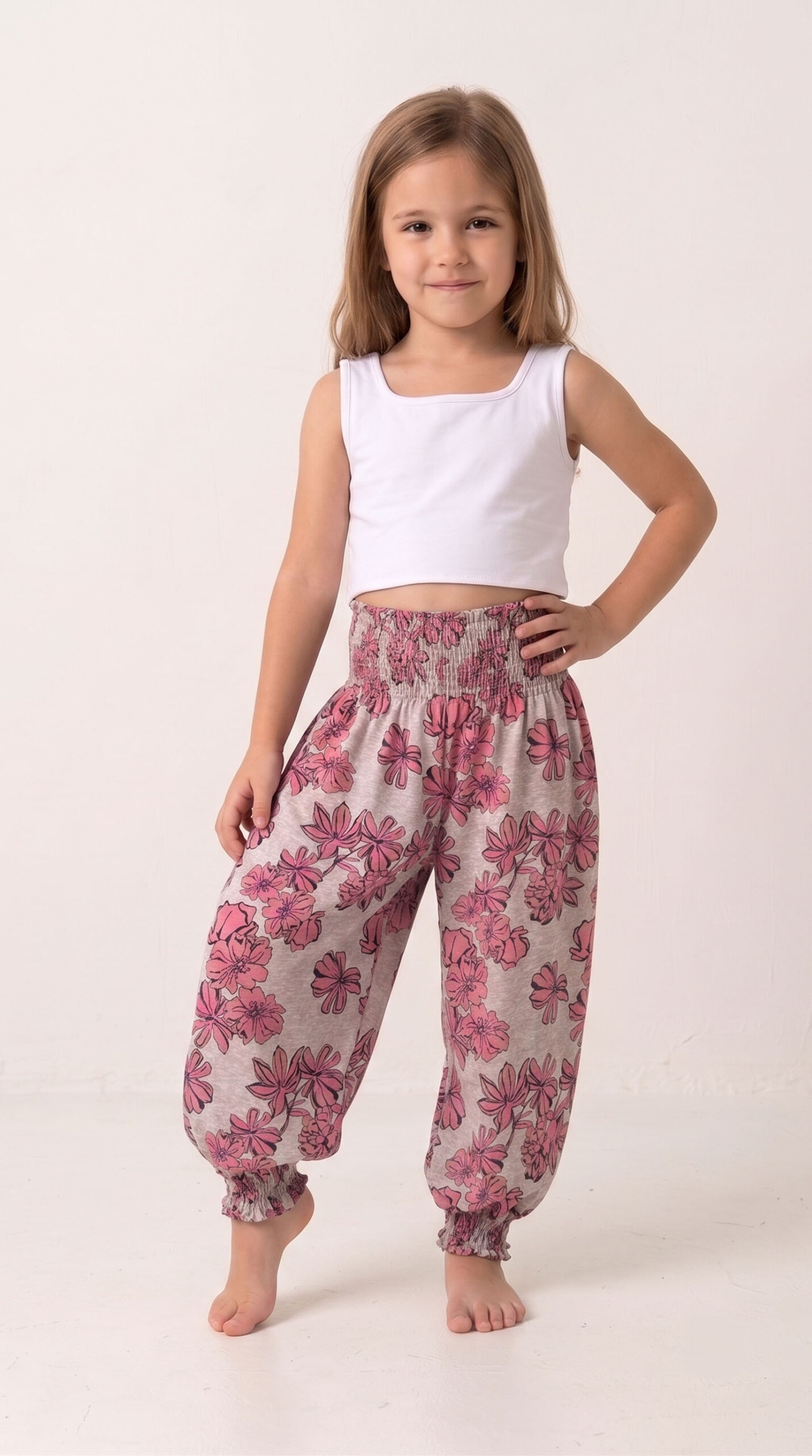 Niña con pelo liso vistiendo un pantalón estilo harem modelo Alicia con estampado de flores rosas sobre fondo claro, cintura y tobillos fruncidos con nido de abeja, combinando con un top blanco.