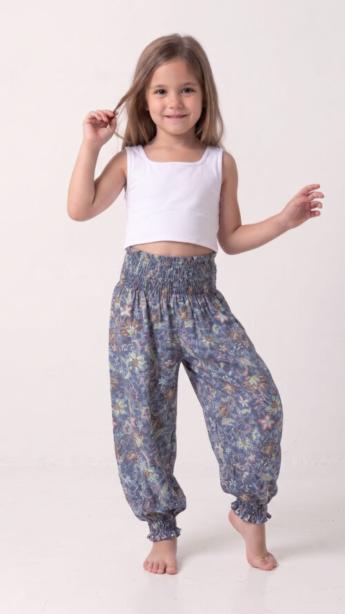 Niña sonriente y descalza posando con el pantalón harem modelo Aurora, que presenta un estampado floral bohemio en tonos tierra sobre azul polvo, combinado con un top blanco básico.
