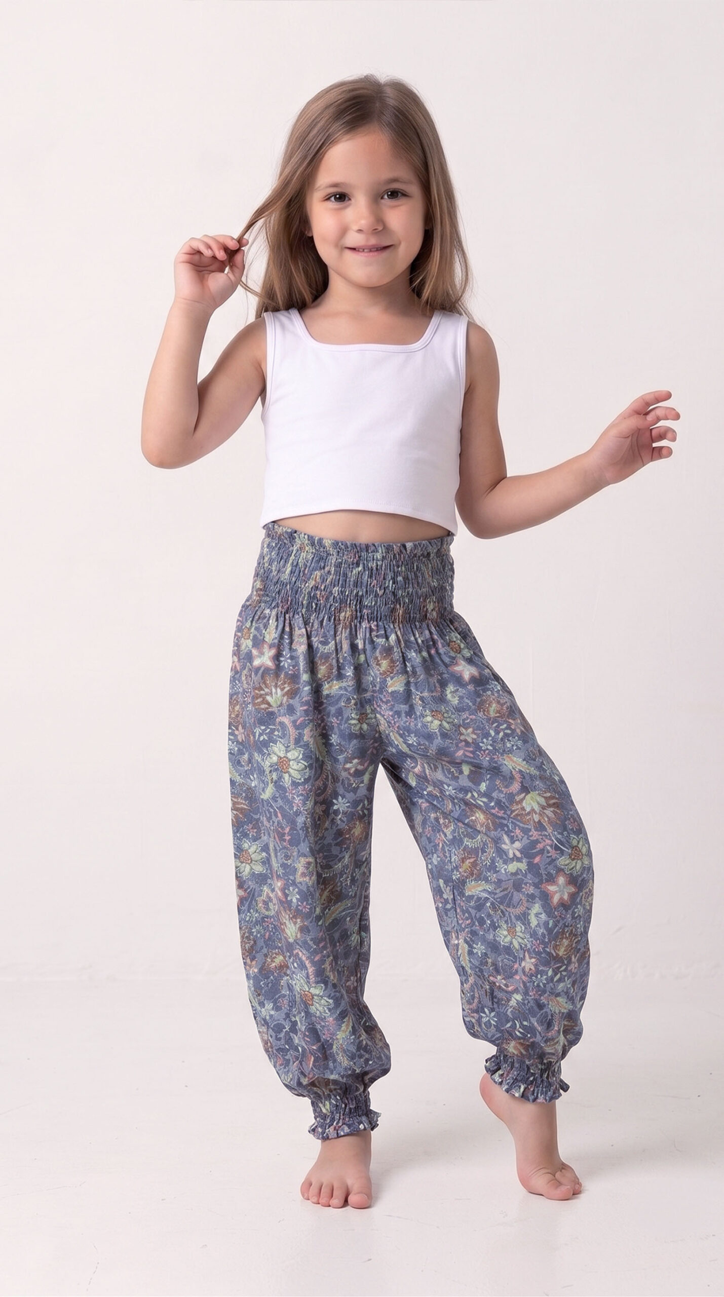 Niña sonriente y descalza posando con el pantalón harem modelo Aurora, que presenta un estampado floral bohemio en tonos tierra sobre azul polvo, combinado con un top blanco básico.