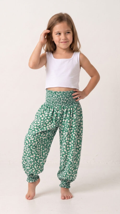 Niña descalza y sonriente posando con un top blanco y el pantalón harem Lara, destacando su vibrante estampado de pequeñas flores blancas sobre fondo verde y su cintura elástica fruncida.