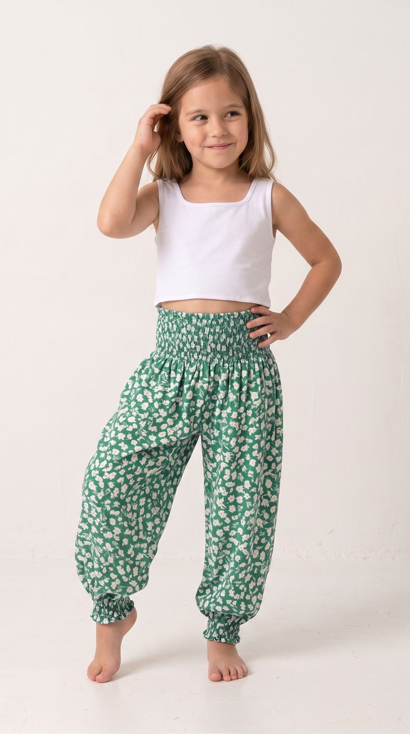 Niña descalza y sonriente posando con un top blanco y el pantalón harem Lara, destacando su vibrante estampado de pequeñas flores blancas sobre fondo verde y su cintura elástica fruncida.