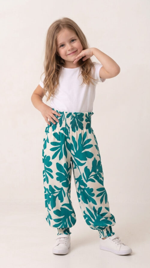 Niña sonriendo vistiendo un pantalón estilo harem modelo Lia con un vibrante estampado de hojas verdes teal sobre fondo blanco, cintura y tobillos fruncidos, combinado con camiseta blanca.