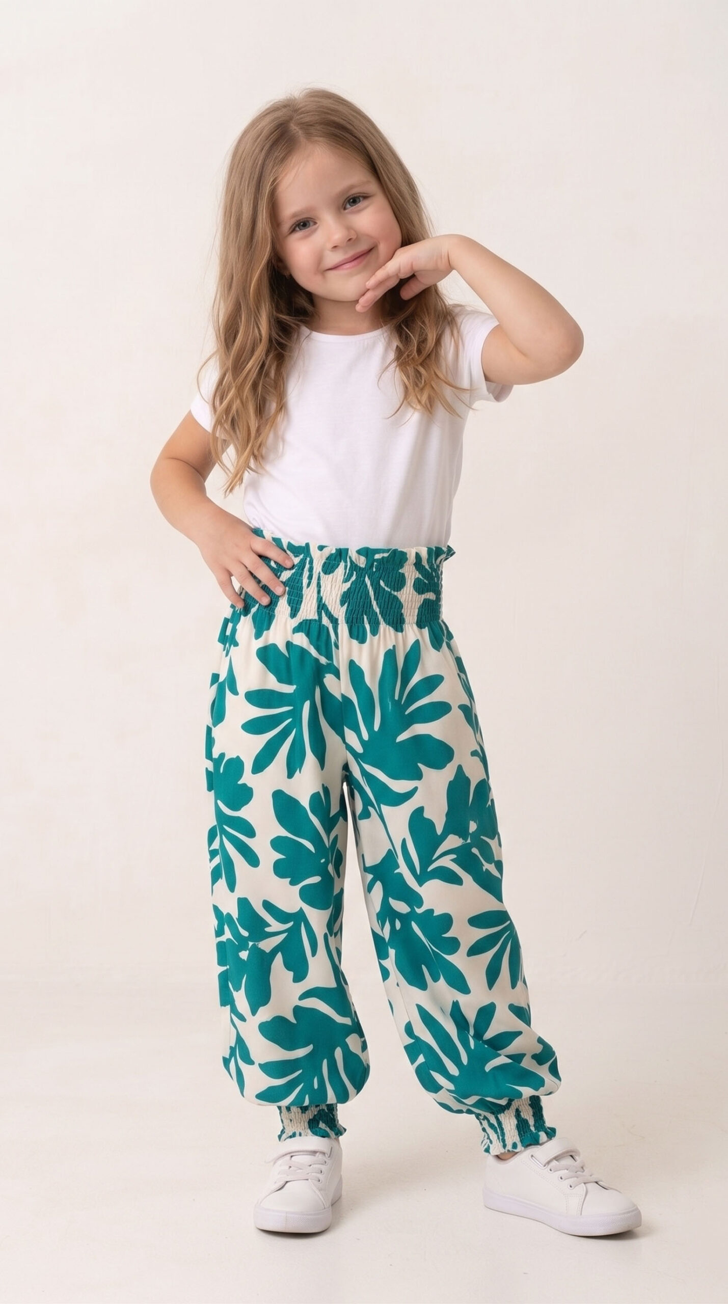 Niña sonriendo vistiendo un pantalón estilo harem modelo Lia con un vibrante estampado de hojas verdes teal sobre fondo blanco, cintura y tobillos fruncidos, combinado con camiseta blanca.